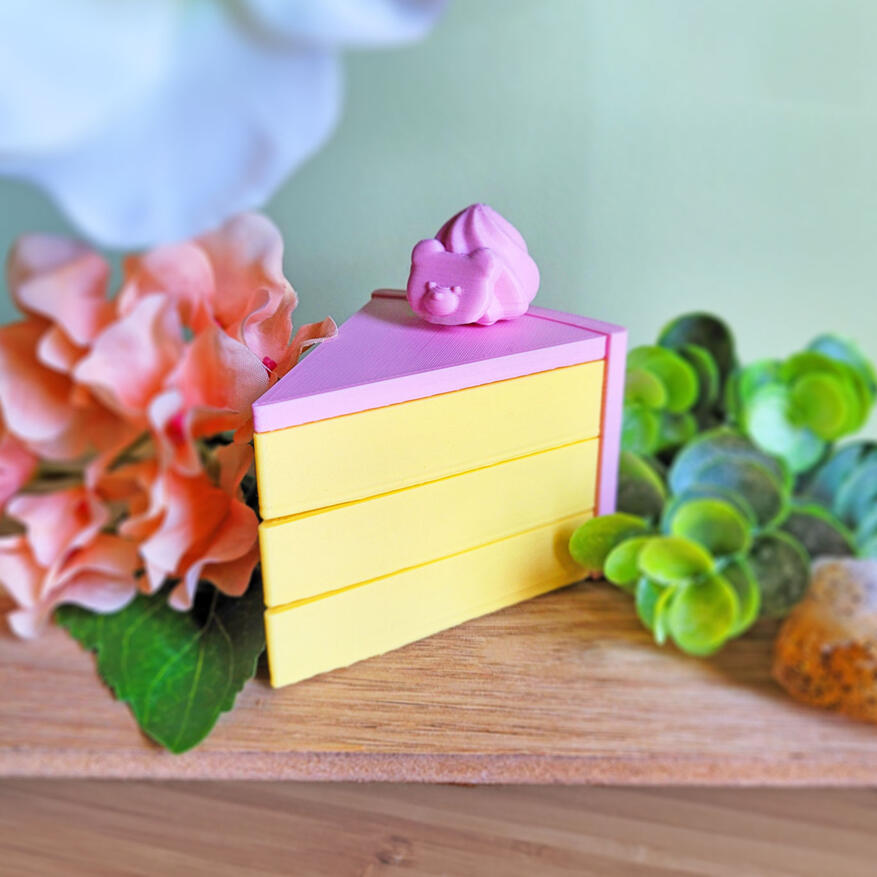 Untitled A pink and yellow 3D printed boxe, modeled to look like a slice of cake. The box has 3 compartments, each one a "layer" of cake. There is a small decoration on the top of it. The box is closed.