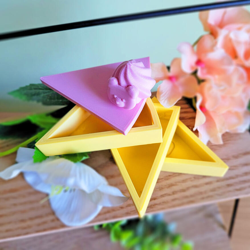 A pink and yellow 3D printed boxe, modeled to look like a slice of cake. The box has 3 compartments, each one a "layer" of cake. There is a small decoration on the top of it. The box is open, showing how all three compartments swivel around an axis.
