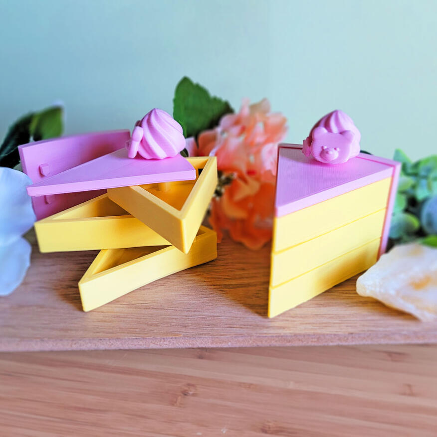 Two pink and yellow 3D printed boxes, modeled to look like slices of cake. The boxes have 3 compartments, each one a "layer" of cake. There is a small decoration on the top of them. One of them is closed, and one of them is open, showing how the compartmen